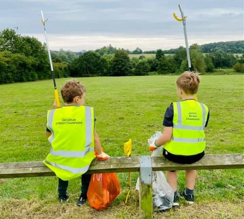 Two boys sat on a fence, facing away from the camera with their litter pickers in the air. They wear hi-vis vests with 'Junior Street Champions' on the back. 